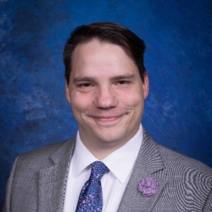 Man in a suit coat and tie smiling at camera with blue background.