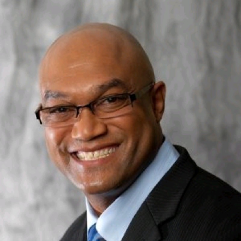 A man in a suit and tie smiles at the camera against a gray mottled background. He is wearing glasses.