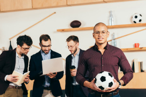 A man holding a soccer ball stands in front of three sports management professionals in a room with modern wooden shelves