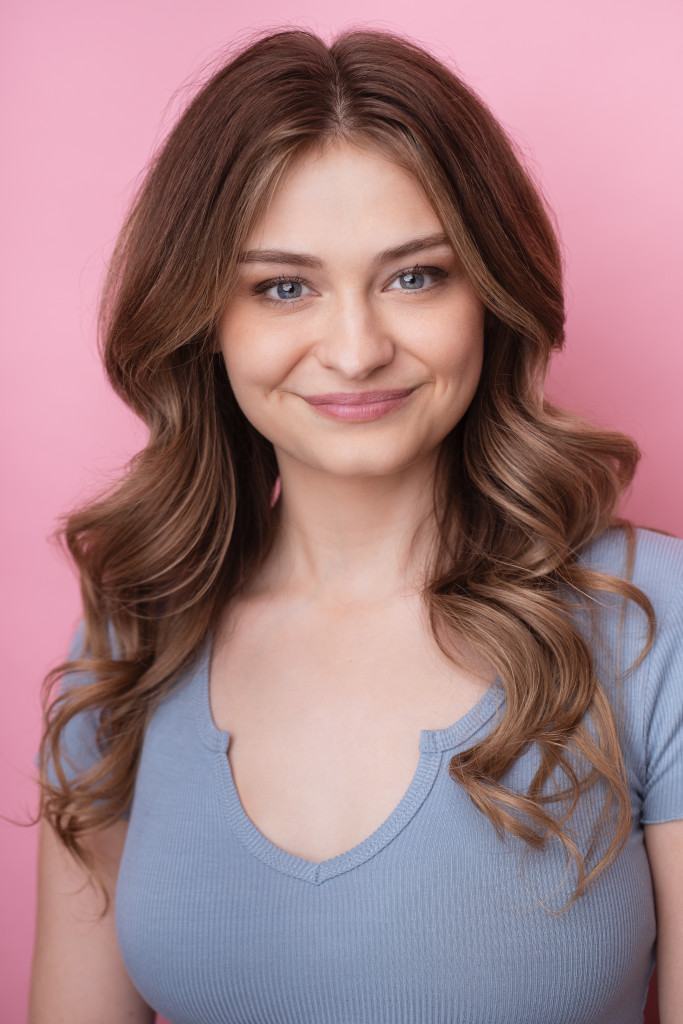 Commercial headshot of a Russell Sage College BA in Theatre graduate, a young woman with wavy brown hair, smiling in front of a pink background and wearing a light blue top.