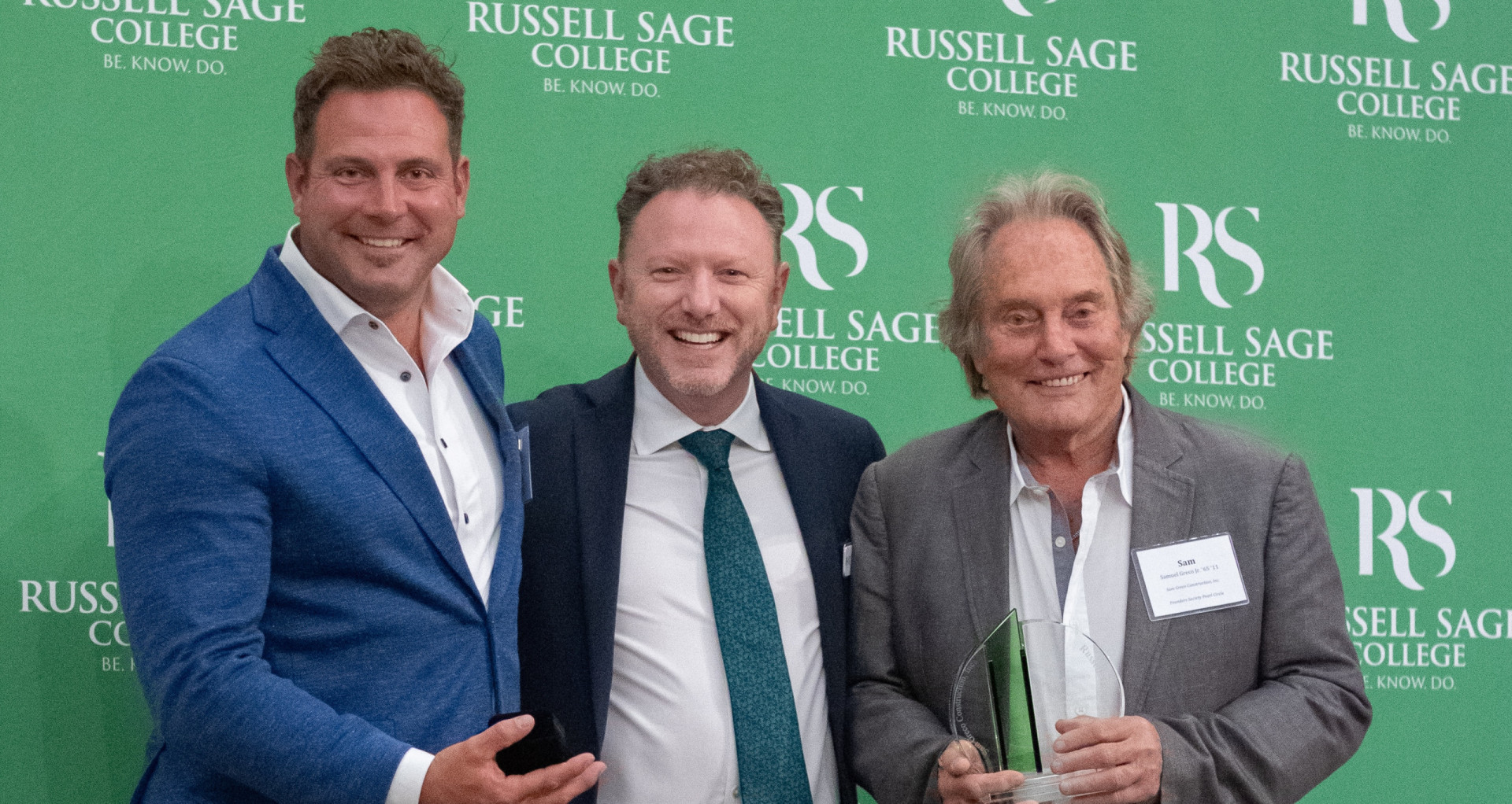 three men dressed in sport coats smiling at the camera in front of a green backdrop that says Russell Sage College.