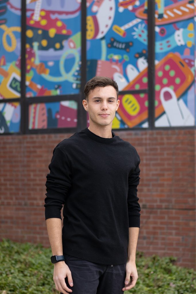 A young man in a black shirt stands in front of a colorful mural with geometric shapes.