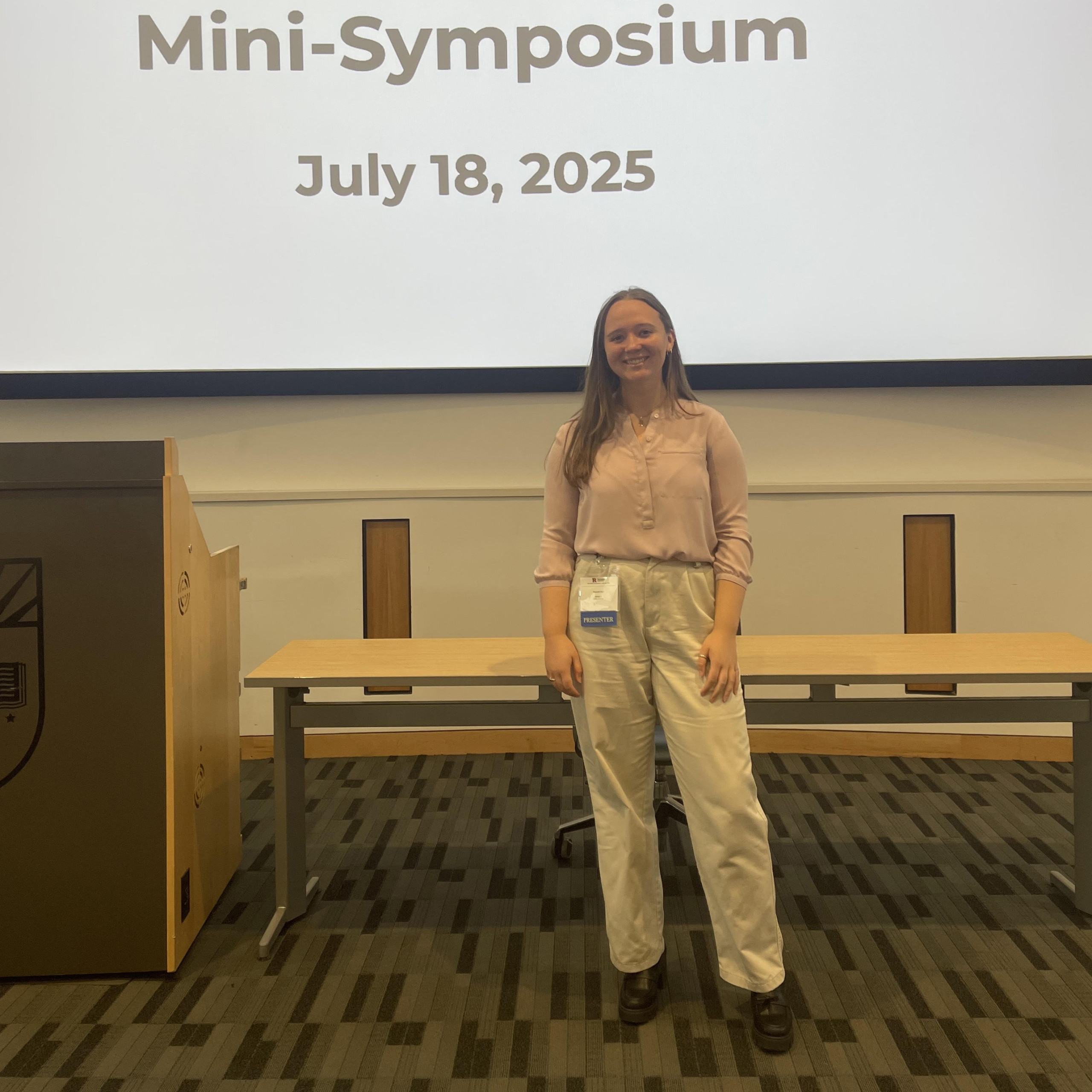 A woman stands confidently in front of a screen displaying "Mini-Symposium, July 18, 2025." She's dressed in a pink blouse and white pants.