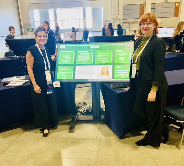 Two women stand beside a green electronic poster display at a conference.