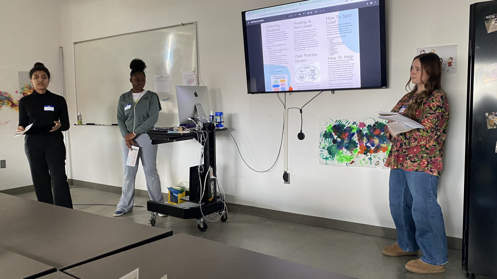 Three individuals presenting in a classroom with a monitor and a whiteboard.