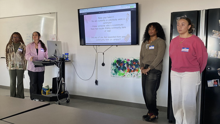 Four people presenting in a classroom with a large screen displaying a slide