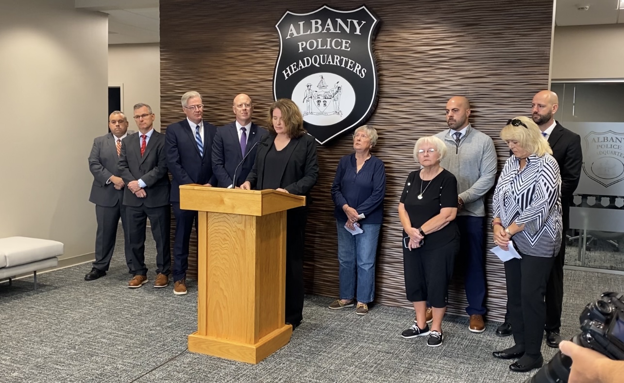 Woman dressed in black suit stands at podium with line of several people standing behind her and an "Albany Police Department" sign on the wall.
