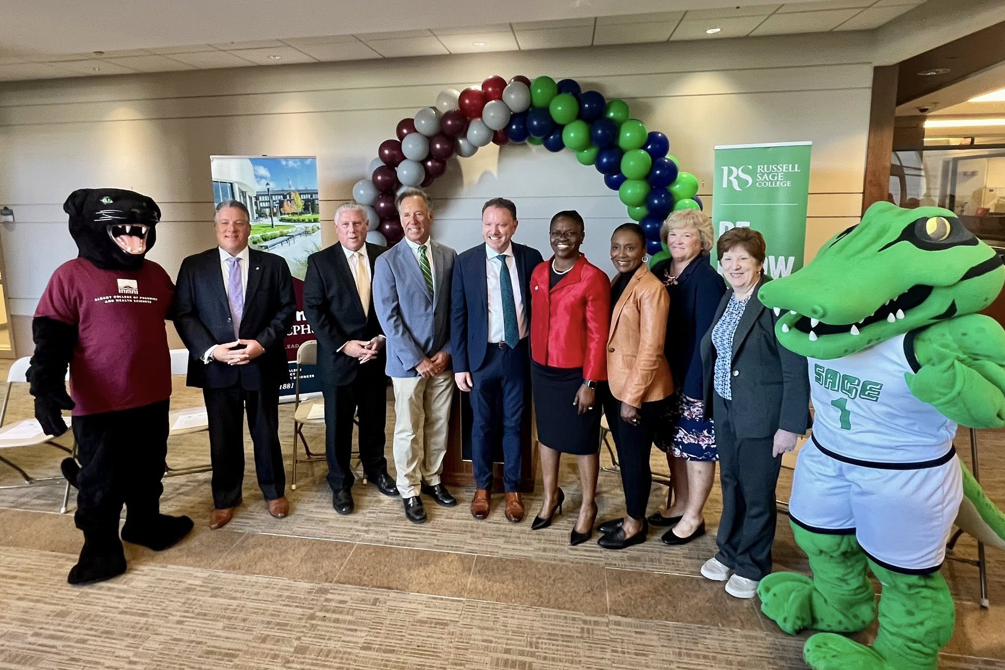 8 people dressed professionally stand smiling at the camera in front of balloon arch, where half of the balloons are maroon and silver and half are blue and green.