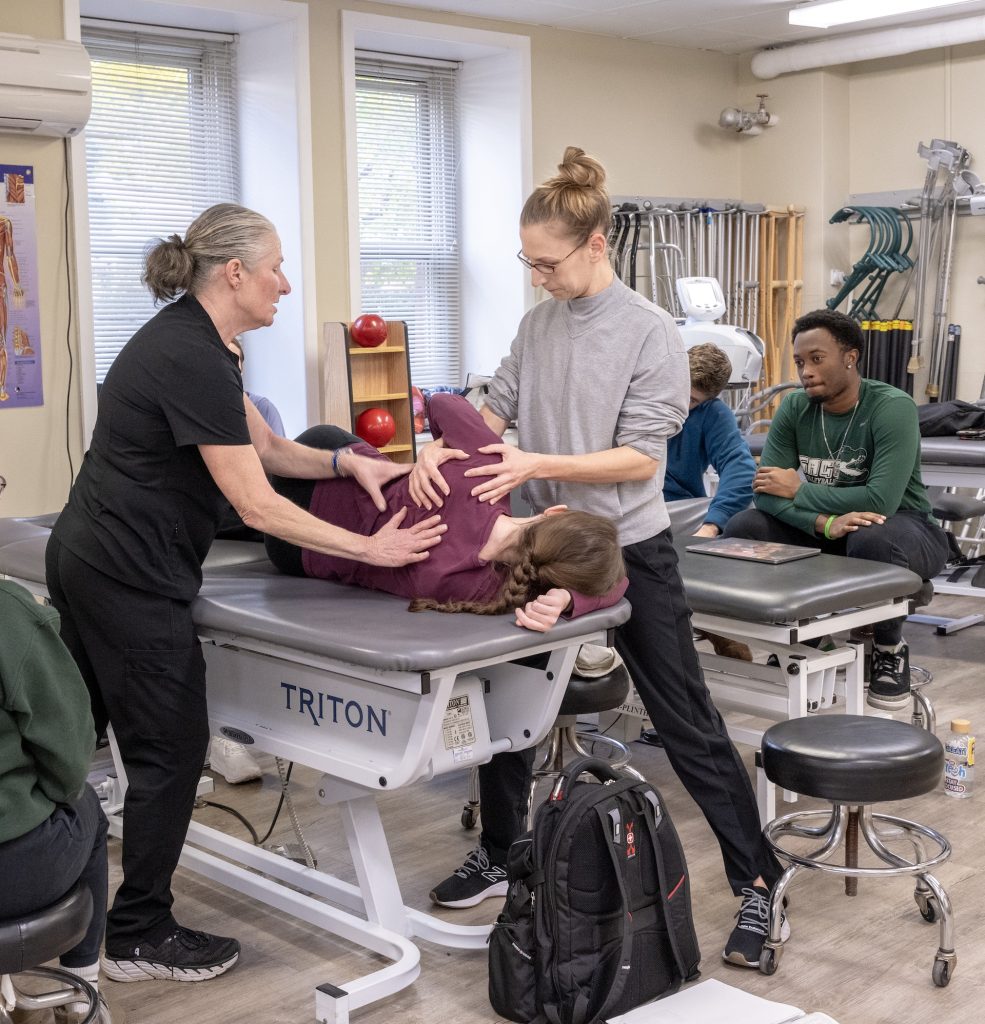 Two people standing around a therapy table working on the shoulder of another person lying on their side.