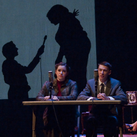 Two people seated at a table with microphones, in front of projected silhouettes on a stage.