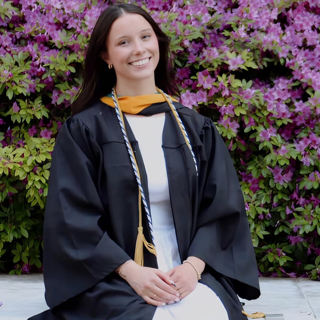 Person in graduation attire sitting in front of flowering bushes.