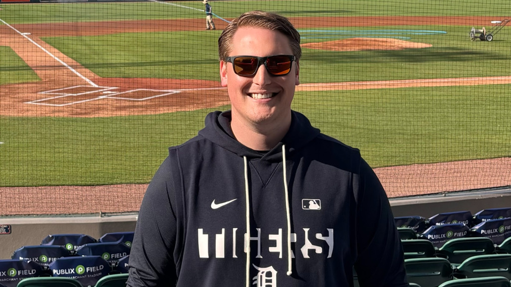Person standing in front of a baseball field, wearing a Detroit Tigers hoodie and sunglasses, with a scoreboard in the background.