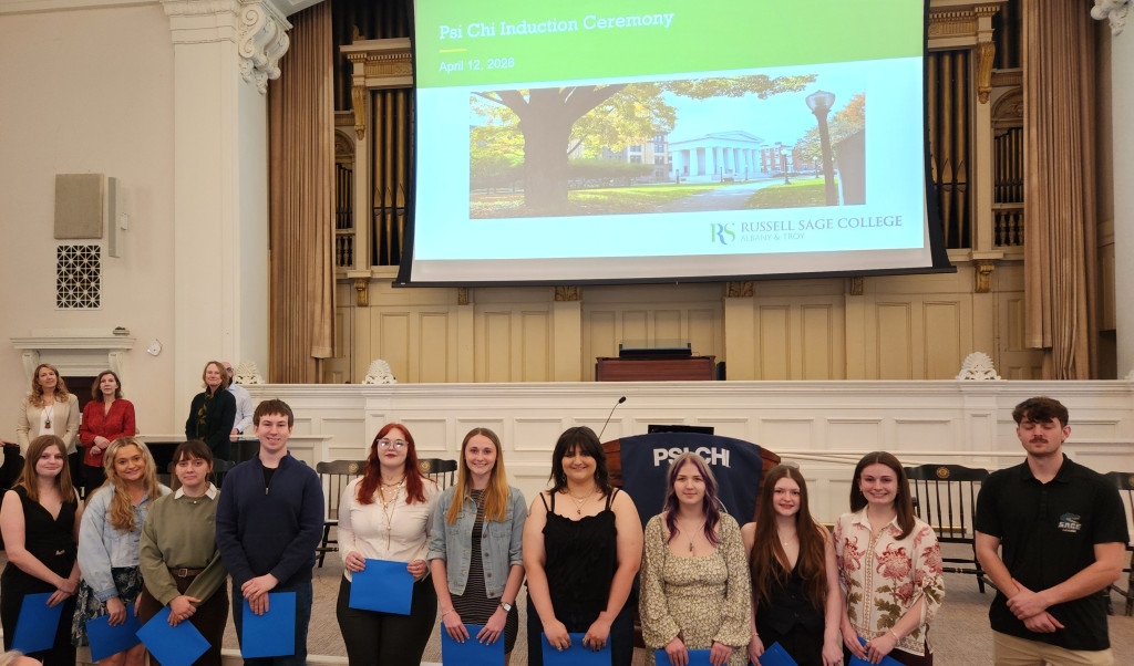 A group of young adults stand holding blue folders at a Psi Chi induction ceremony in a hall with a display screen above showing event details.