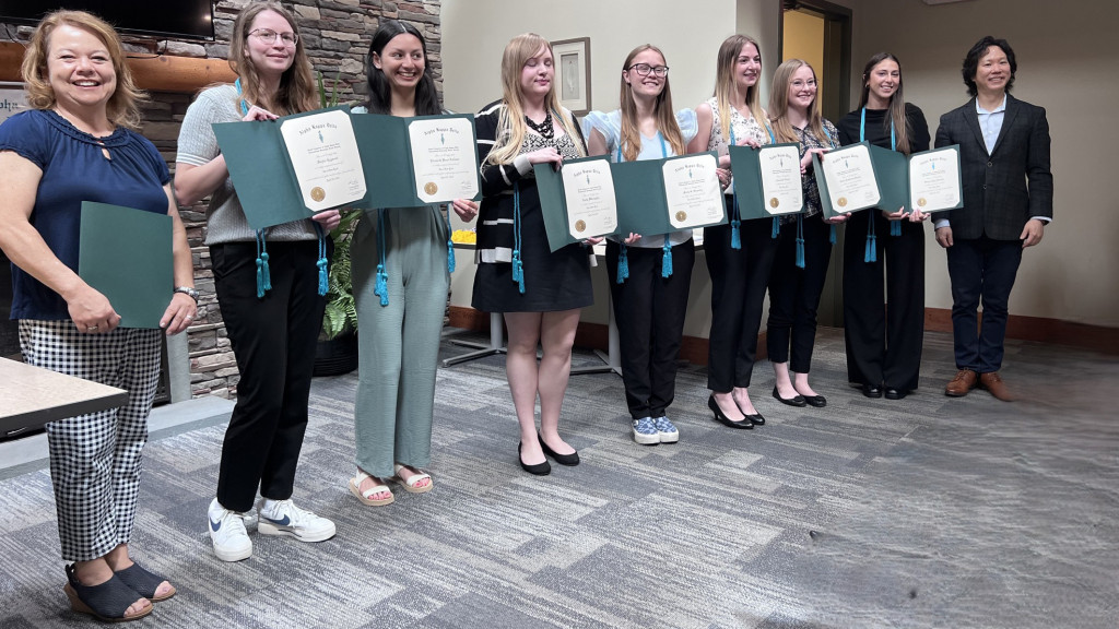 Nine people standing in a line, eight holding certificates in green folders, and smiling indoors.