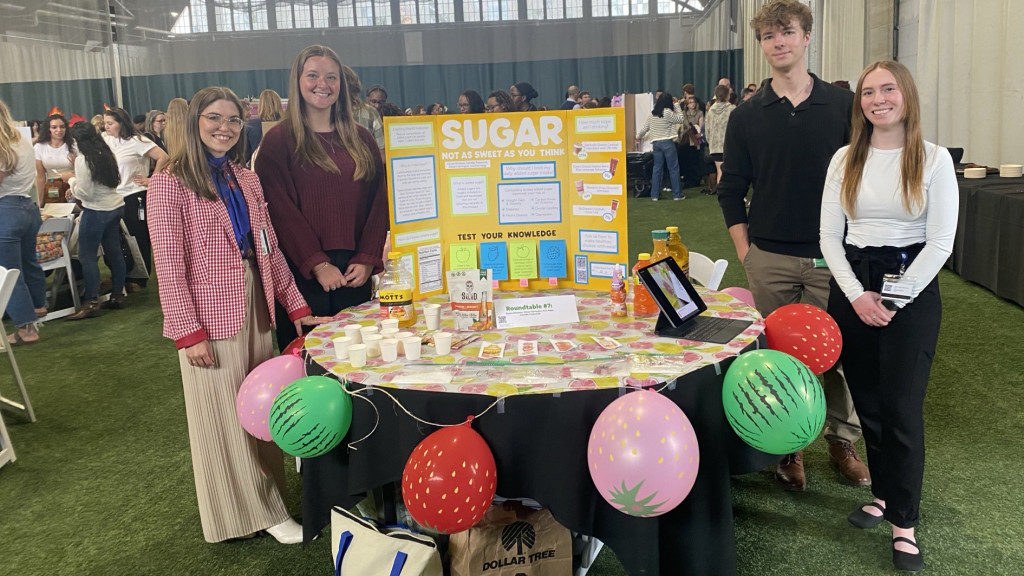 People standing by a display about sugar at an event, with informational board and props on a table.