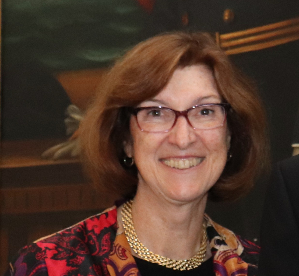 A smiling woman with glasses and shoulder-length brown hair wears a vibrant, patterned blazer and a gold necklace. The background is blurred and indoors.