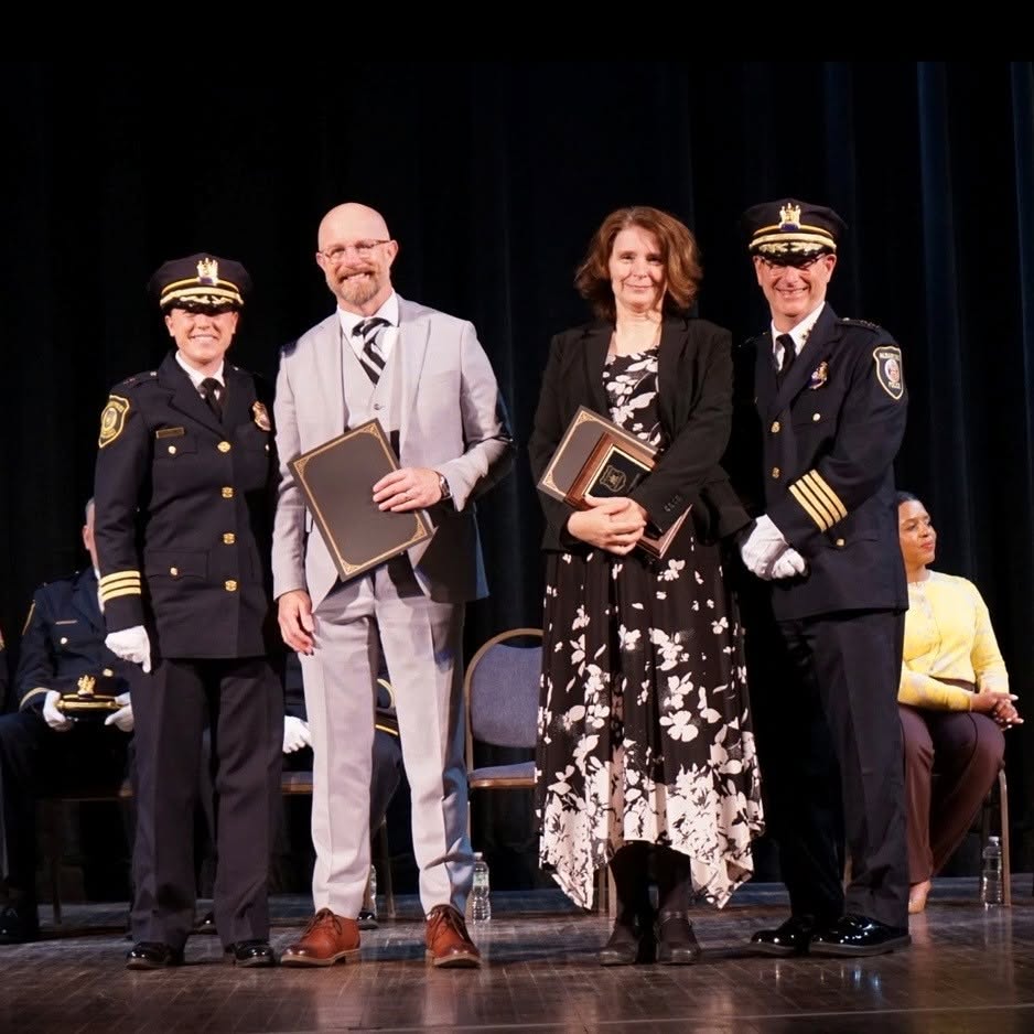 Four people standing in a row on a stage with awards. The man and woman in the center are dressed in professional office clothing, on either side of them are police officers in uniform.