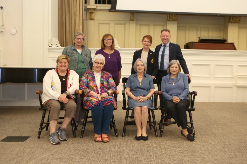 7 women and one man in two rows -- half sitting, half standing -- smiling indoors
