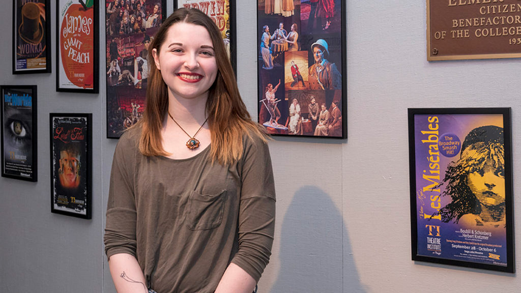 Young woman smiling in front of theater posters, wearing a gray long-sleeve shirt and blue jeans.