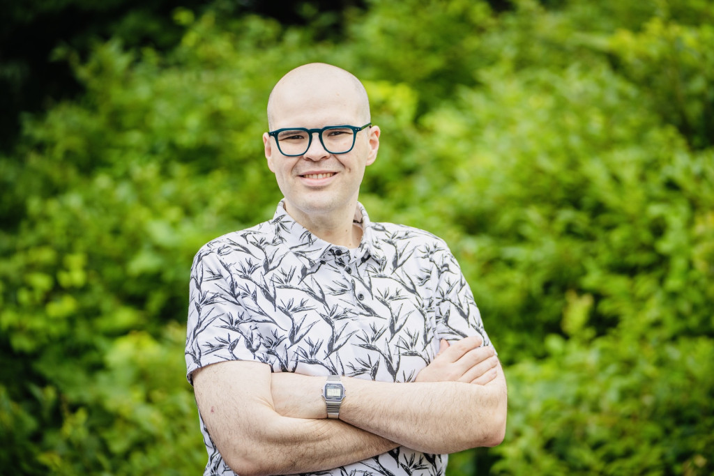 Smiling man wearing glasses and a botanical-print shirt, standing with folded arms against a green leafy background.