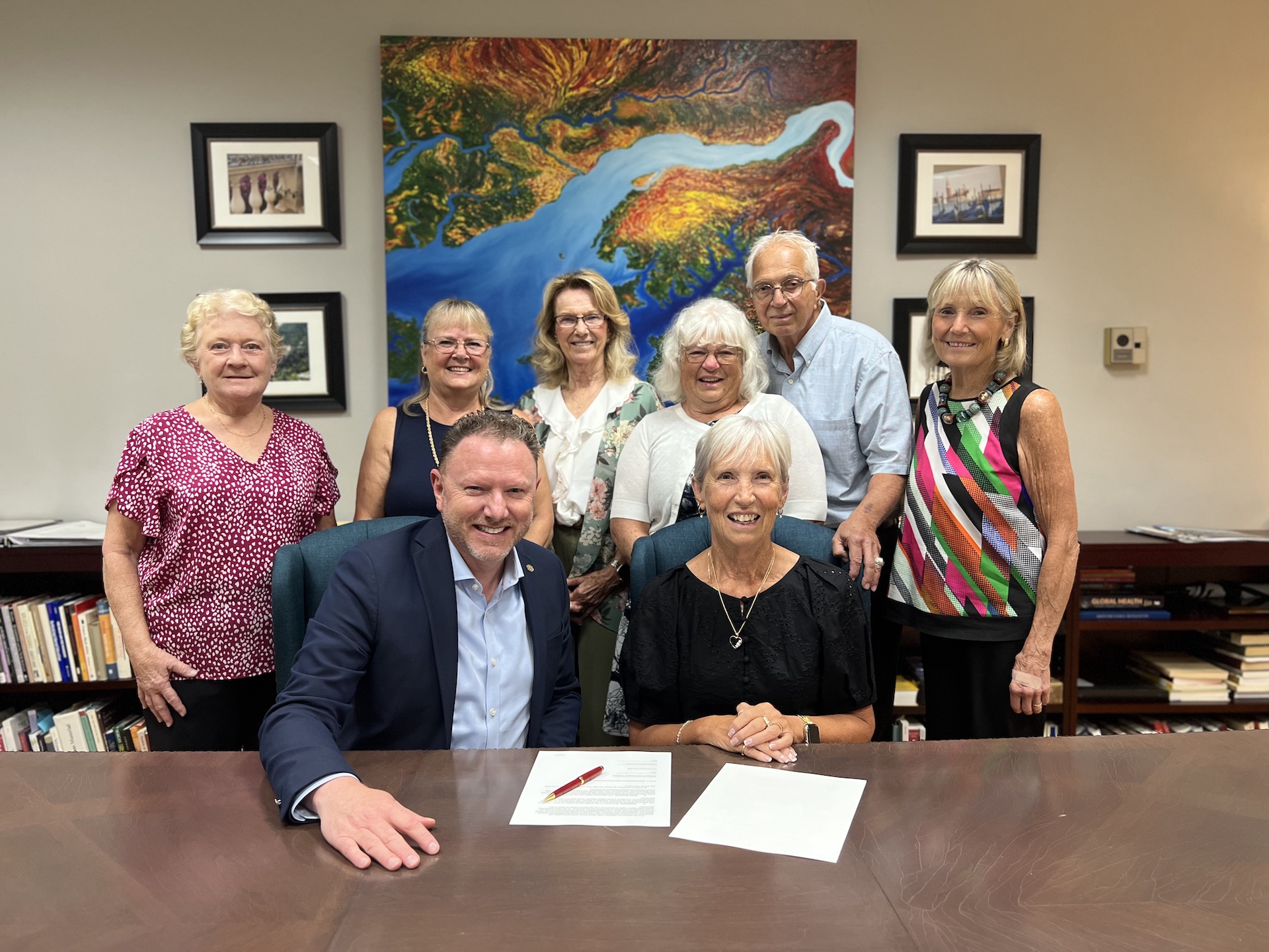 Eight people smiling and looking at the camera and posing for a photo in an office. Two of the people are seated at a table and the rest are standing behind them. There are two pieces of paper and a pen on the table.