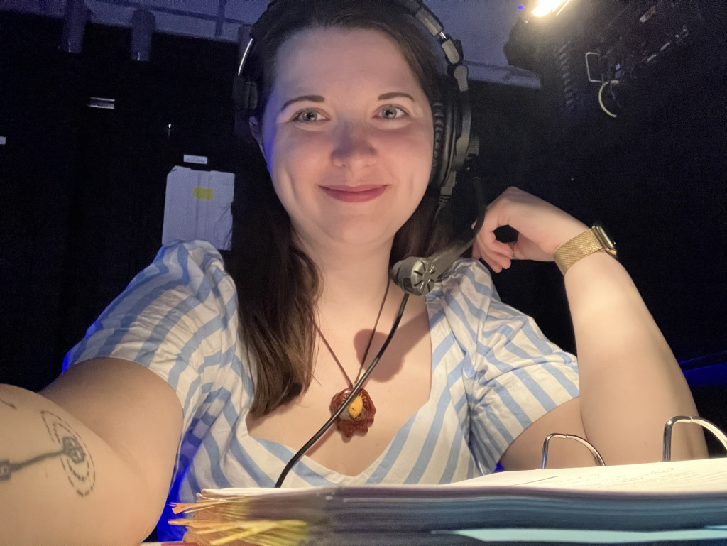 Alt-text: A woman wearing a striped blouse and headset smiles at the camera in a dimly lit room, with papers in front of her.