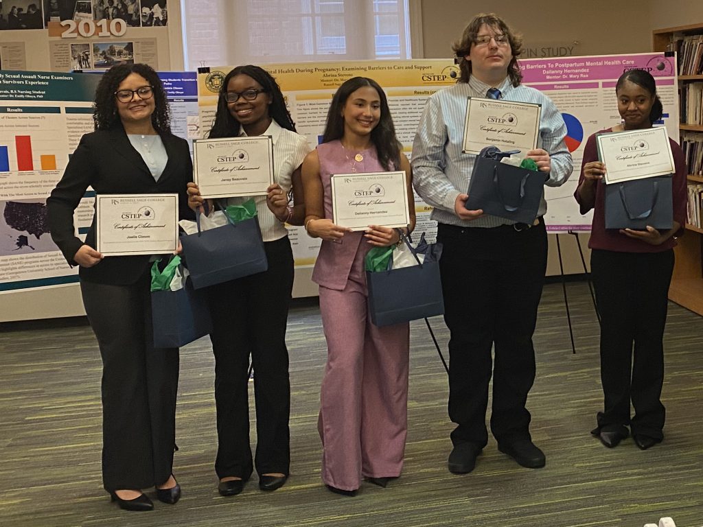 Five individuals stand together holding certificates of accomplishment at an event at Russell Sage College, surrounded by informational posters.