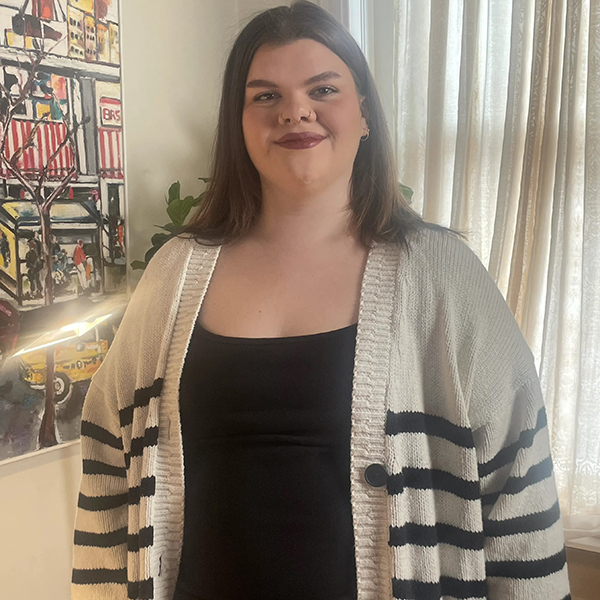A BS in Musical Theatre student with long brown hair, wearing a black top and striped white cardigan, stands indoors near a window and painting.