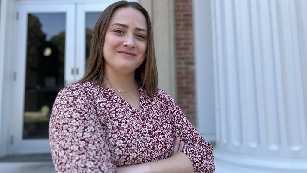 Smiling woman in a burgundy floral blouse stands with arms crossed outside a building on a college campus.
