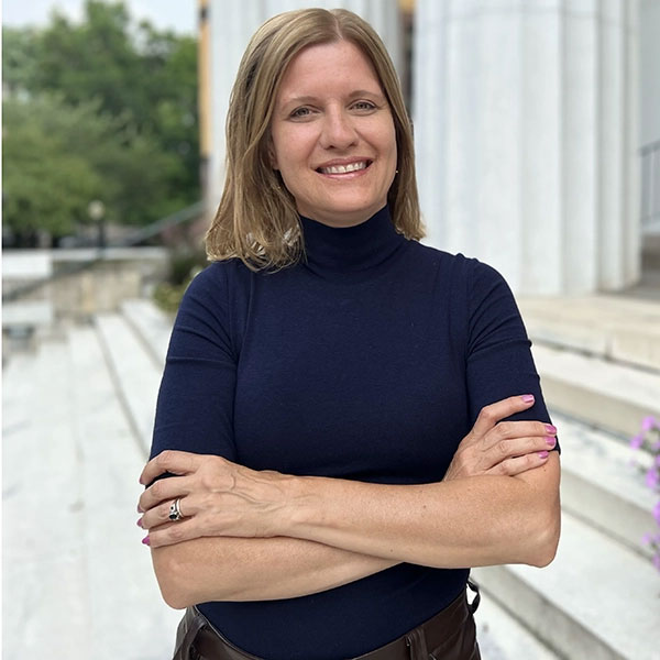 Smiling woman with shoulder-length light hair stands outdoors on steps in a navy turtleneck, arms crossed.