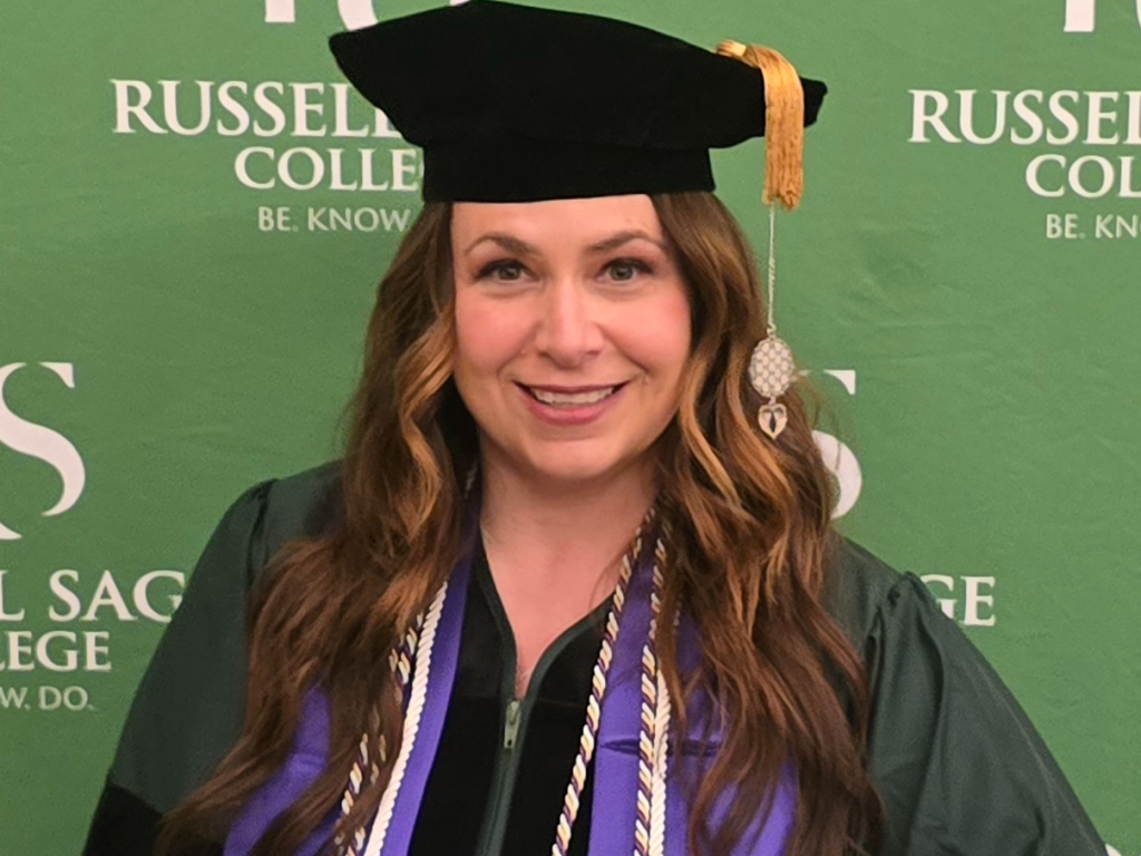 Smiling Ed.D. graduate in black doctoral regalia with a gold tassel and purple-and-white honor cords, posed before a green Russell Sage College backdrop.