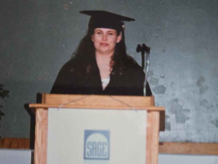 Graduate in black academic regalia standing behind a podium labeled “SAGE JCA.”