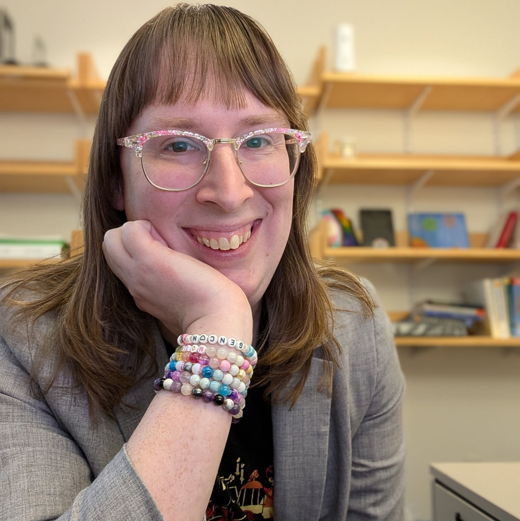 Smiling person with brown hair, pink floral glasses, and beaded bracelets resting chin on hand in front of bookshelves.