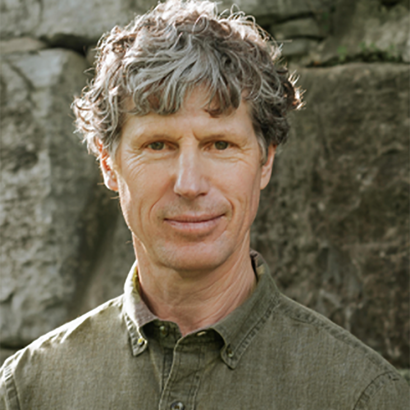 Close-up outdoor portrait of a middle-aged man with tousled gray-brown hair and a slight smile, wearing an olive-green shirt.