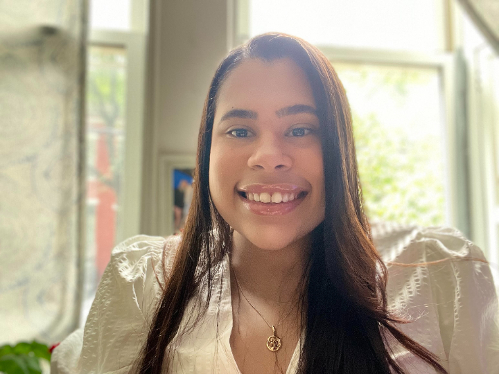 Smiling person with long dark hair wearing a white blouse and pendant necklace, photographed indoors in front of sunlit windows.