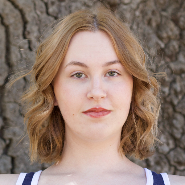 Close-up portrait of a young woman with wavy light-brown hair standing in front of tree bark.