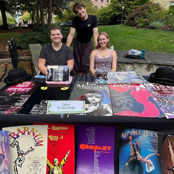 Three people pose behind an outdoor theater display table covered with musical posters and a “Gator Productions” sign.