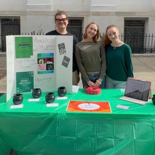 Three people promote a theater program at a table displaying audition flyers, black containers, a QR code, and a sign reading “Auditions Still Open!”