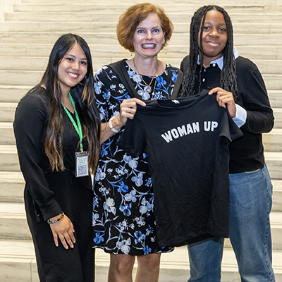 Three people smile while holding a black T-shirt reading “WOMAN UP.”