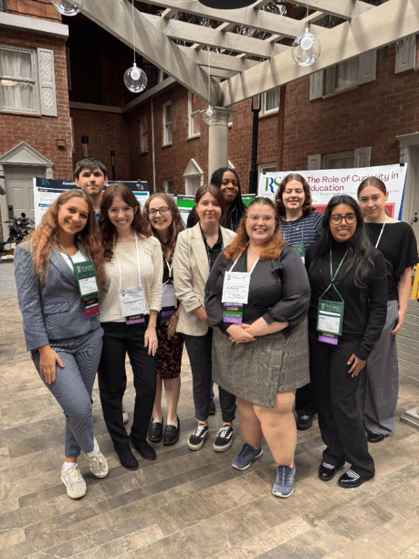 Ten conference attendees pose together beneath a glass-roofed courtyard, with academic poster boards behind them.